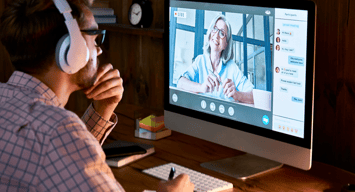 Man wearing headphones over his ears sitting at his computer taking notes, taking an online course with a female instructor.